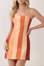 A woman modeling a sleeveless mini dress with vertical color-blocked stripes in terracotta red, orange, and light peach against a plain light beige studio background.