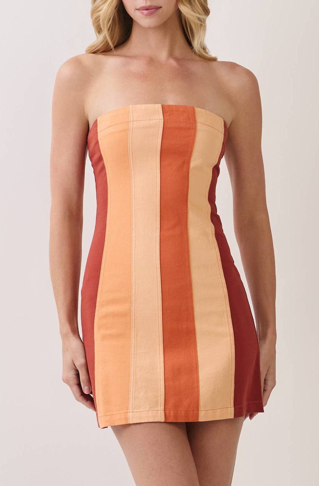 A woman modeling a strapless mini dress with vertical stripes in orange, beige, and red, paired with clear high-heeled sandals, against a plain light beige studio background.