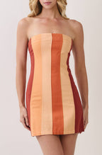 A woman modeling a strapless mini dress with vertical stripes in orange, beige, and red, paired with clear high-heeled sandals, against a plain light beige studio background.