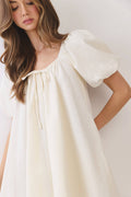 A woman wearing a cream-colored dress with short puff sleeves, a square neckline, and a drawstring detail at the chest, photographed against a plain light background.