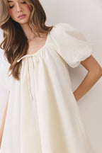 A woman wearing a cream-colored dress with short puff sleeves, a square neckline, and a drawstring detail at the chest, photographed against a plain light background.