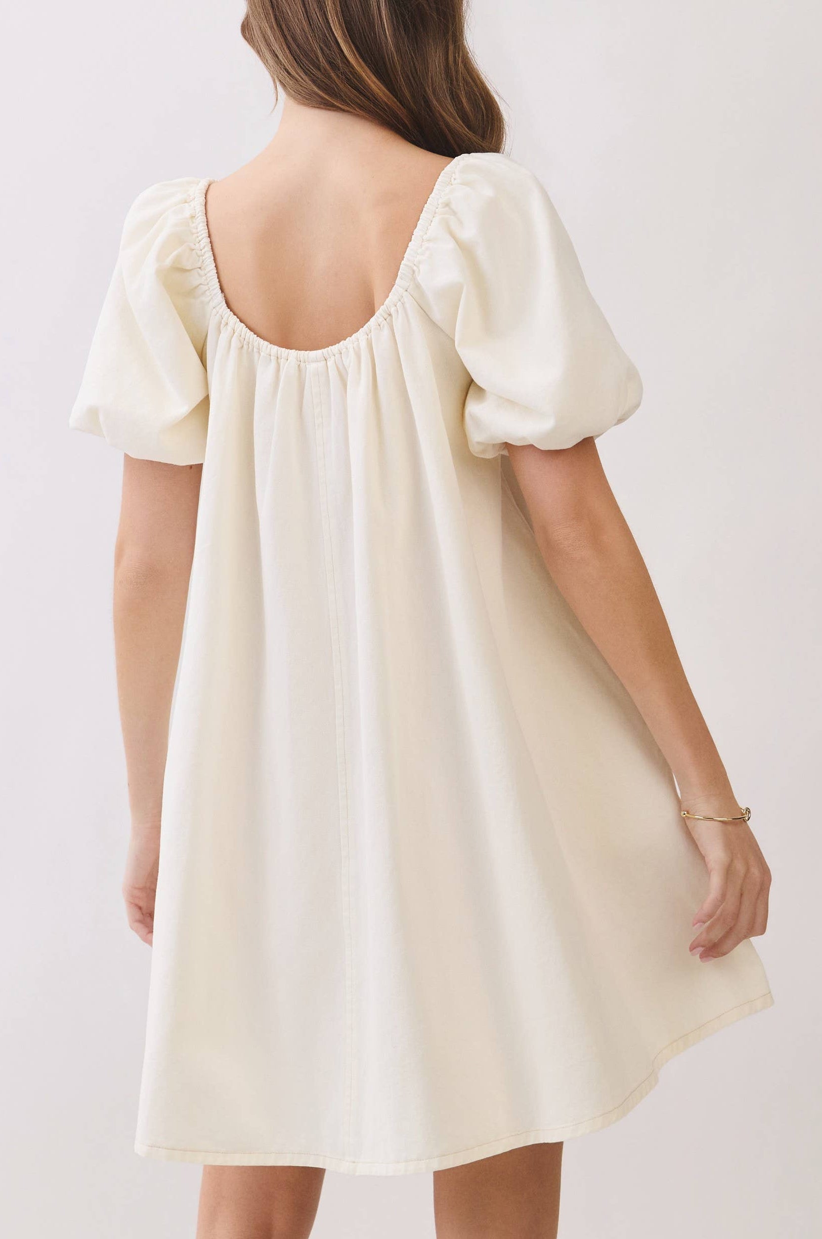 A woman shown from the back wearing a cream-colored puff-sleeve dress against a plain light background.