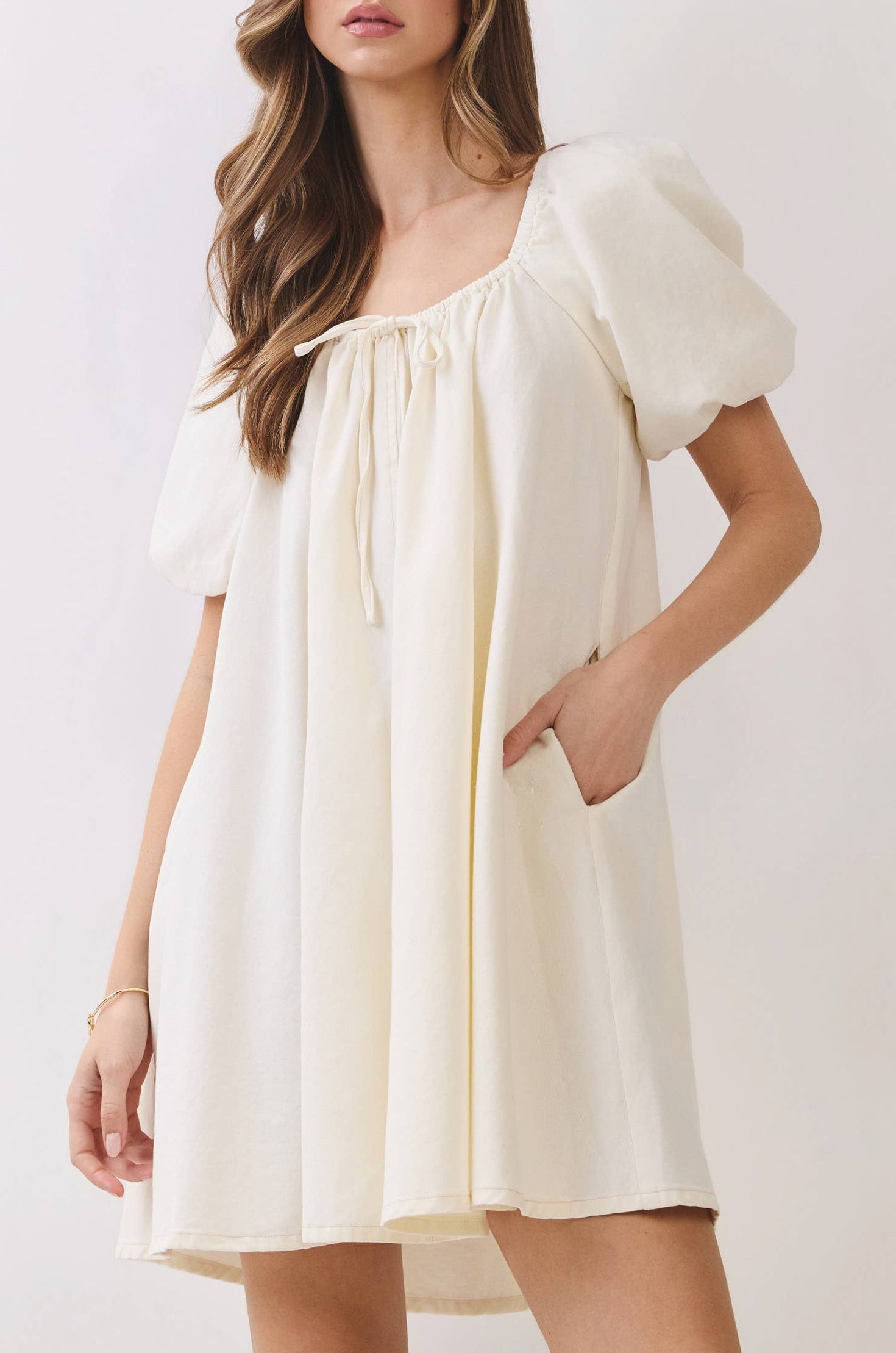 A woman modeling a cream-colored mini dress with puff sleeves, a square neckline with a tie detail, and side pockets, set against a plain light background.