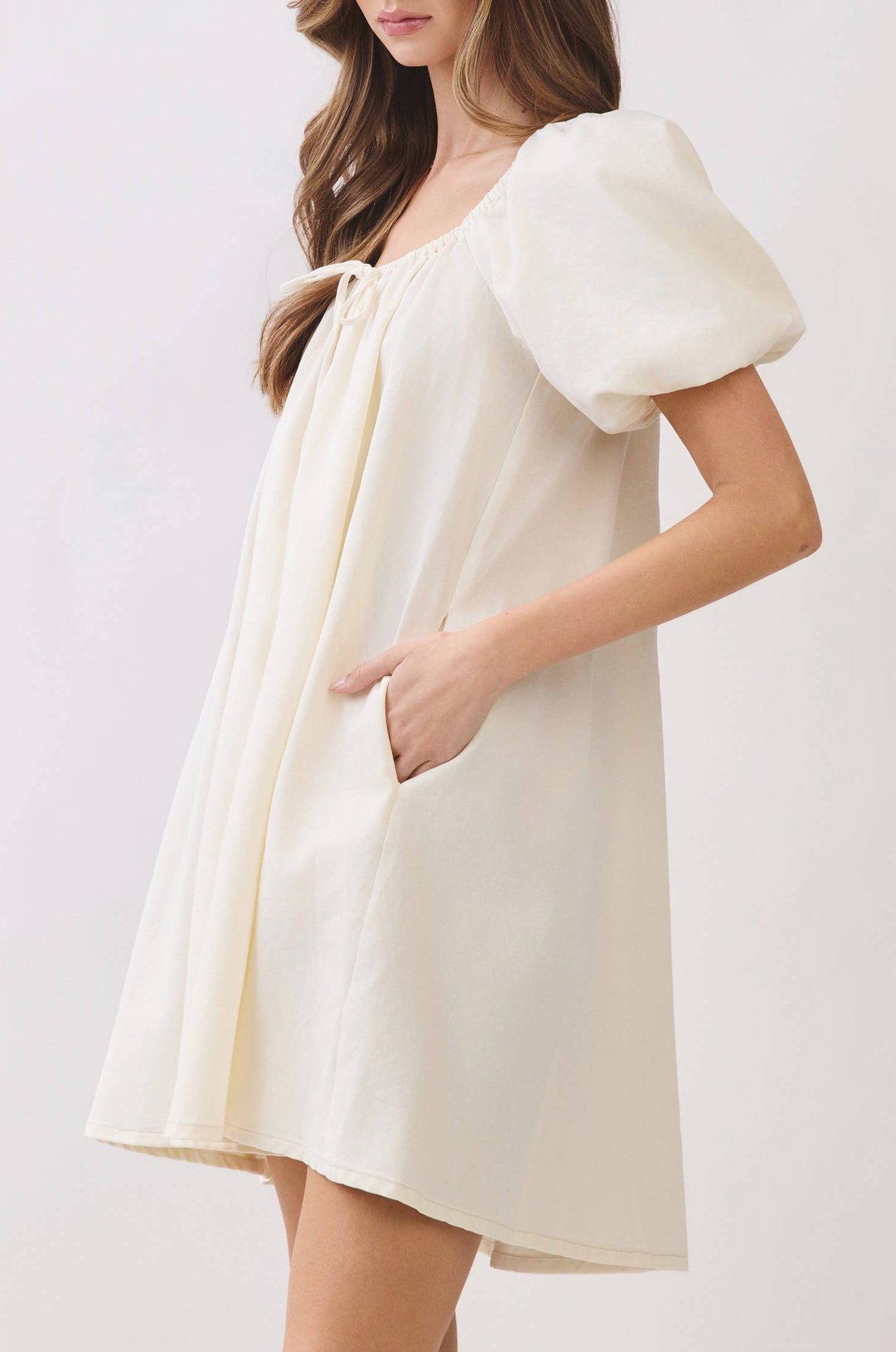 A woman modeling a cream-colored mini dress with puffed short sleeves, a tie neckline, and side pockets, set against a plain light gray studio background.