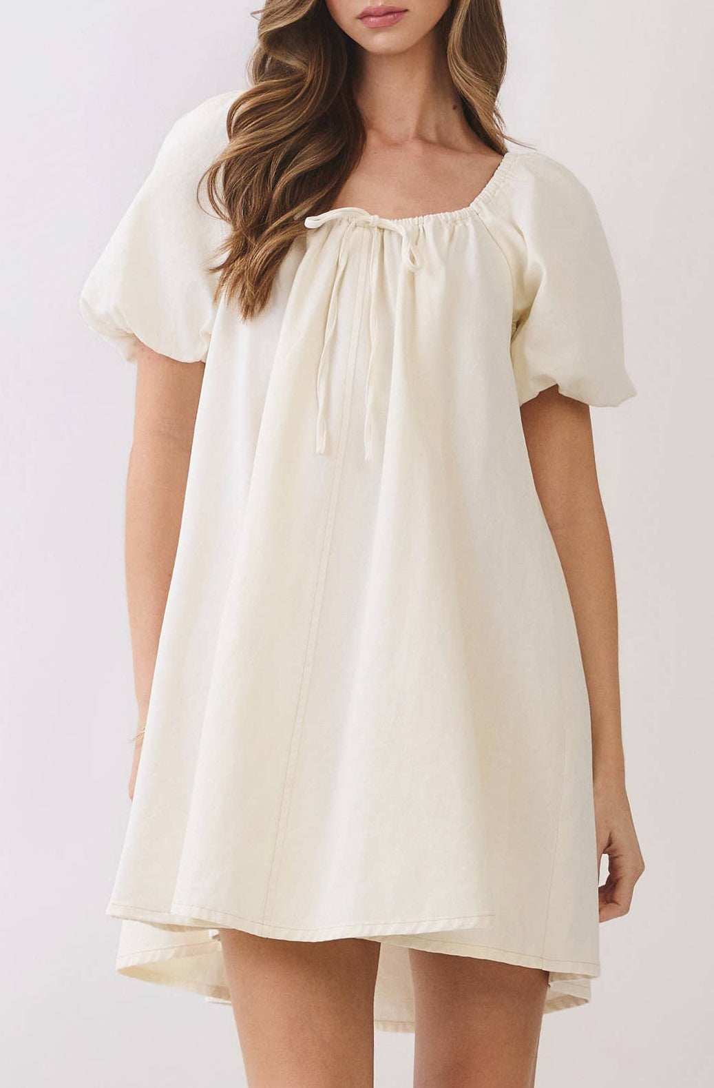 A woman modeling a short cream-colored dress with short puff sleeves, a square neckline with a drawstring, and a loose, flowy silhouette, paired with beige heeled sandals against a light gray studio background.