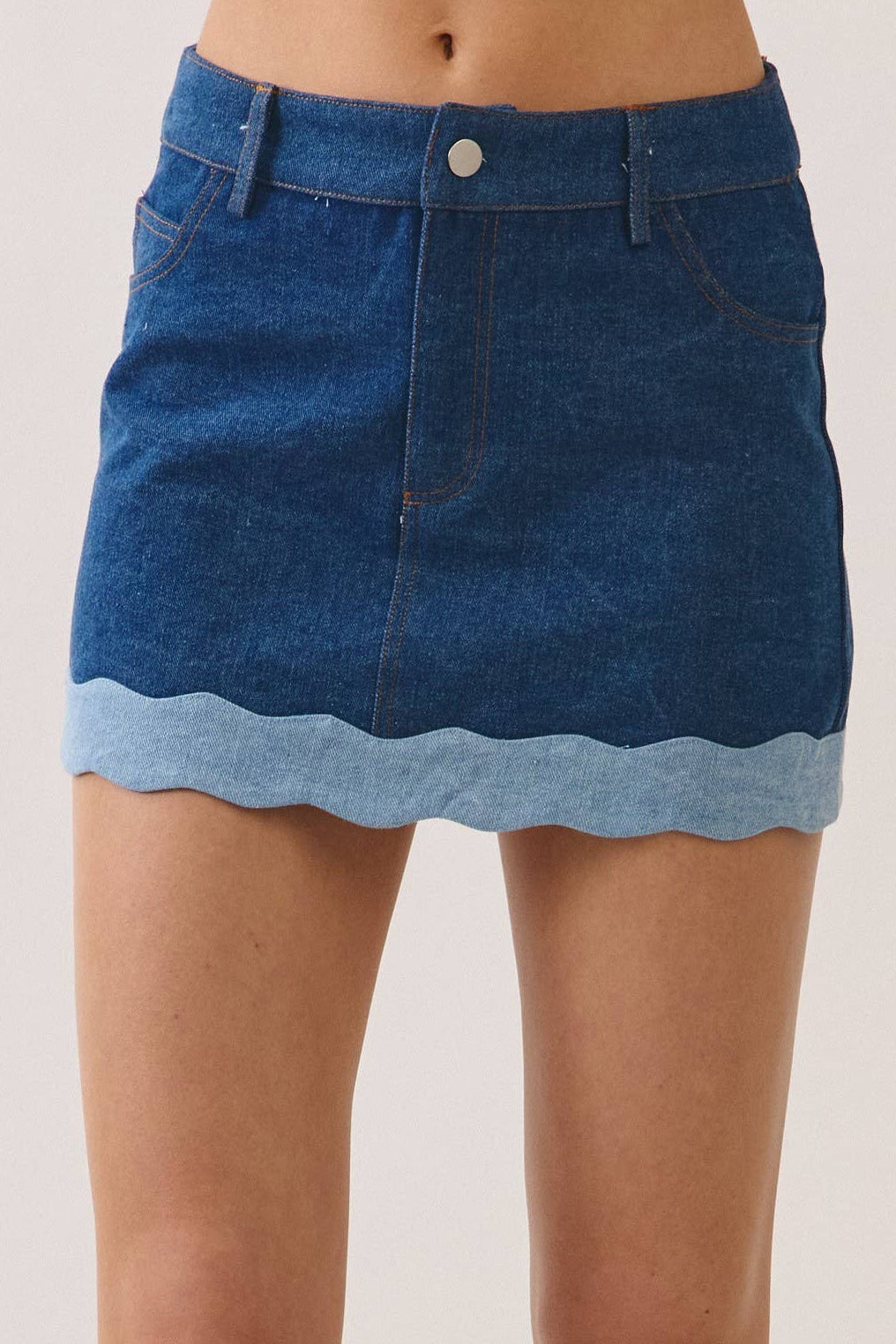 A model's lower body wearing a blue denim mini skirt with a scalloped hem and black sneakers, photographed against a plain light beige studio background.
