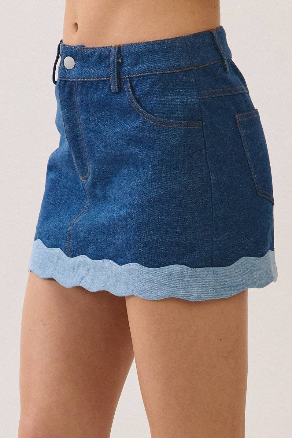 A person wearing a blue denim mini skirt with a scalloped light blue hem, paired with black and white sneakers, shown from the waist down against a plain light beige studio background.