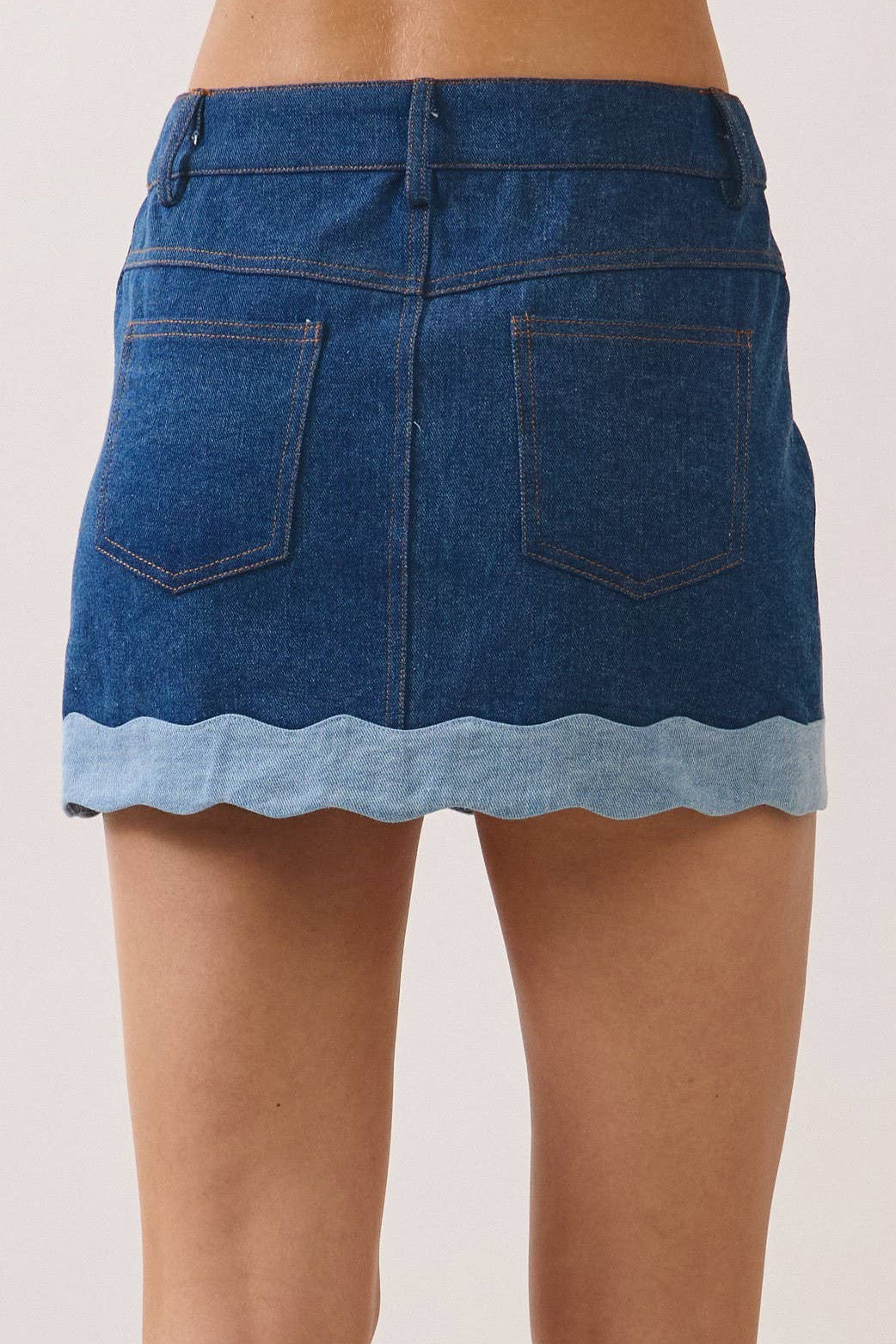 A person shown from the back wearing a blue denim mini skirt with a scalloped light blue hem, paired with black and white sneakers, against a plain light beige studio background.