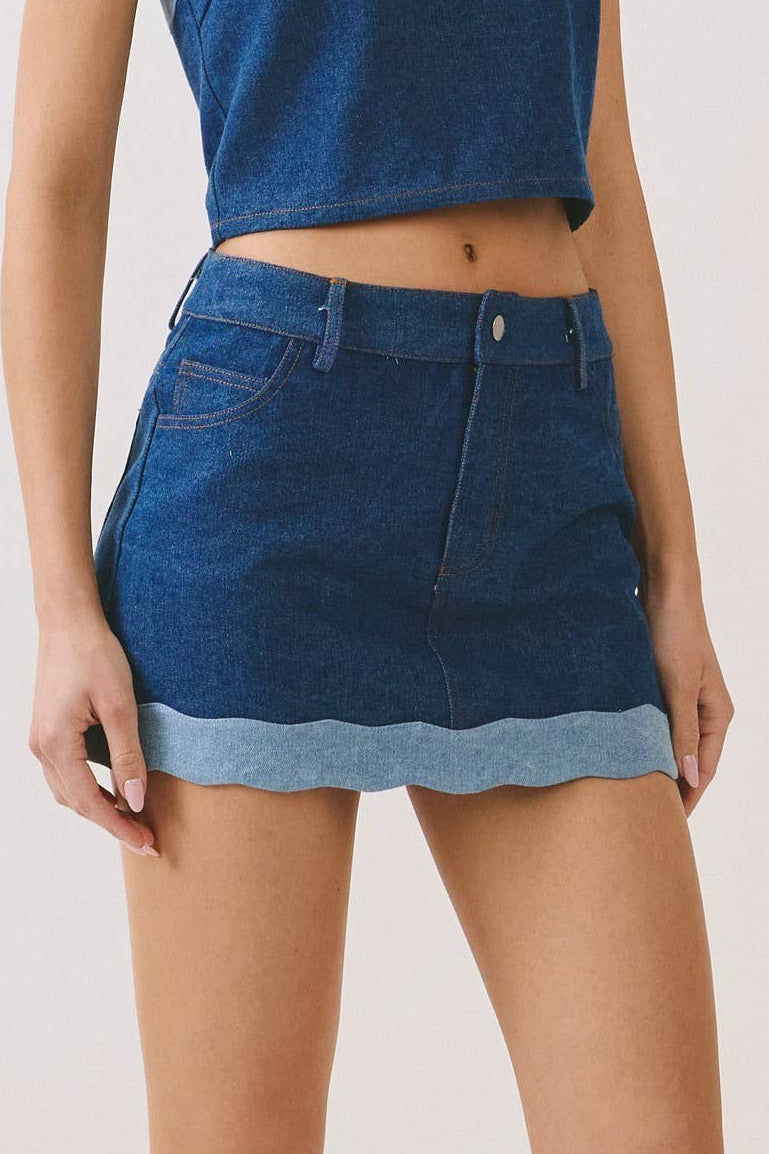 A model wearing a blue denim halter crop top and matching denim mini skirt with light blue contrast trim, paired with black sneakers, against a plain light gray studio background.