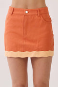An orange denim mini skirt with a scalloped light beige hem, modeled on a person wearing beige heeled sandals against a light gray studio background.