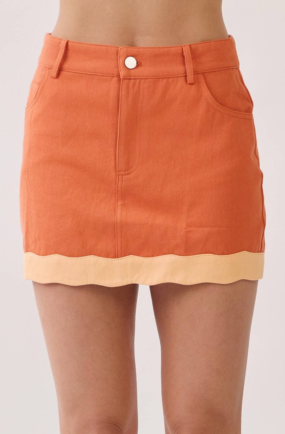 An orange denim mini skirt with a scalloped light beige hem, modeled on a person wearing beige heeled sandals against a light gray studio background.
