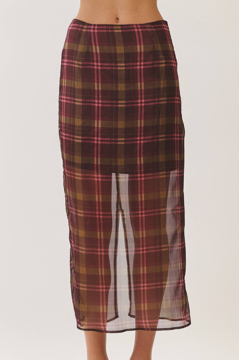 A model wearing a brown and red plaid midi skirt with a sheer front slit, paired with beige heeled sandals, against a plain light background.