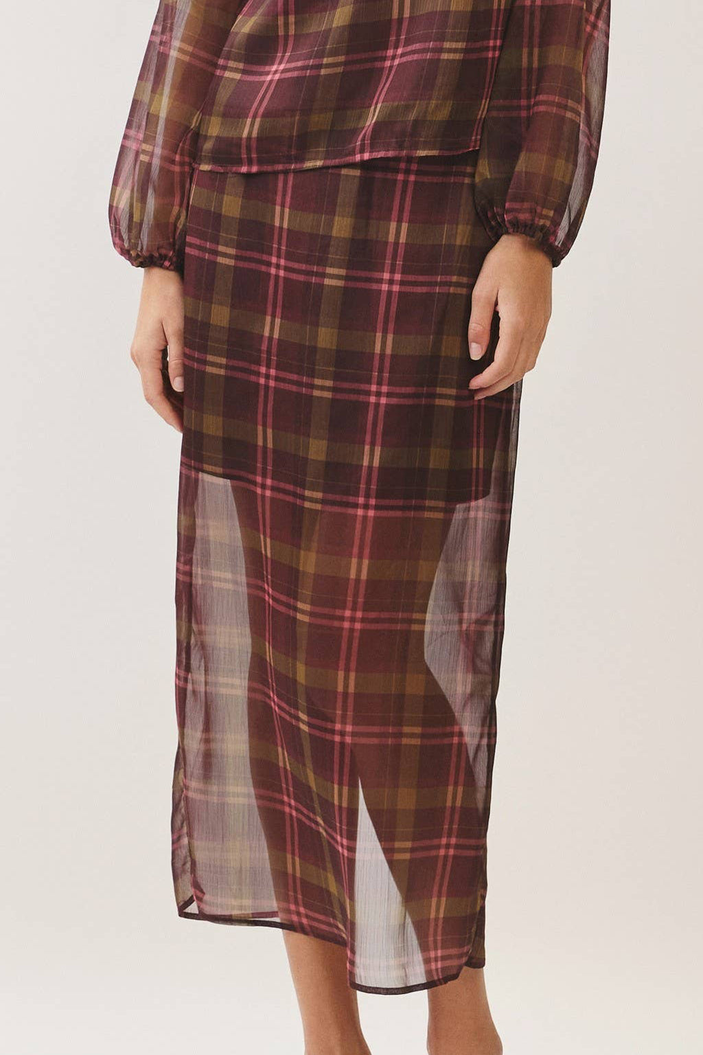 A woman modeling a matching plaid long-sleeve blouse and midi skirt set in a studio setting. The fabric features a brown, red, and black tartan pattern with sheer side panels.