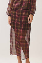 A woman modeling a matching plaid long-sleeve blouse and midi skirt set in a studio setting. The fabric features a brown, red, and black tartan pattern with sheer side panels.