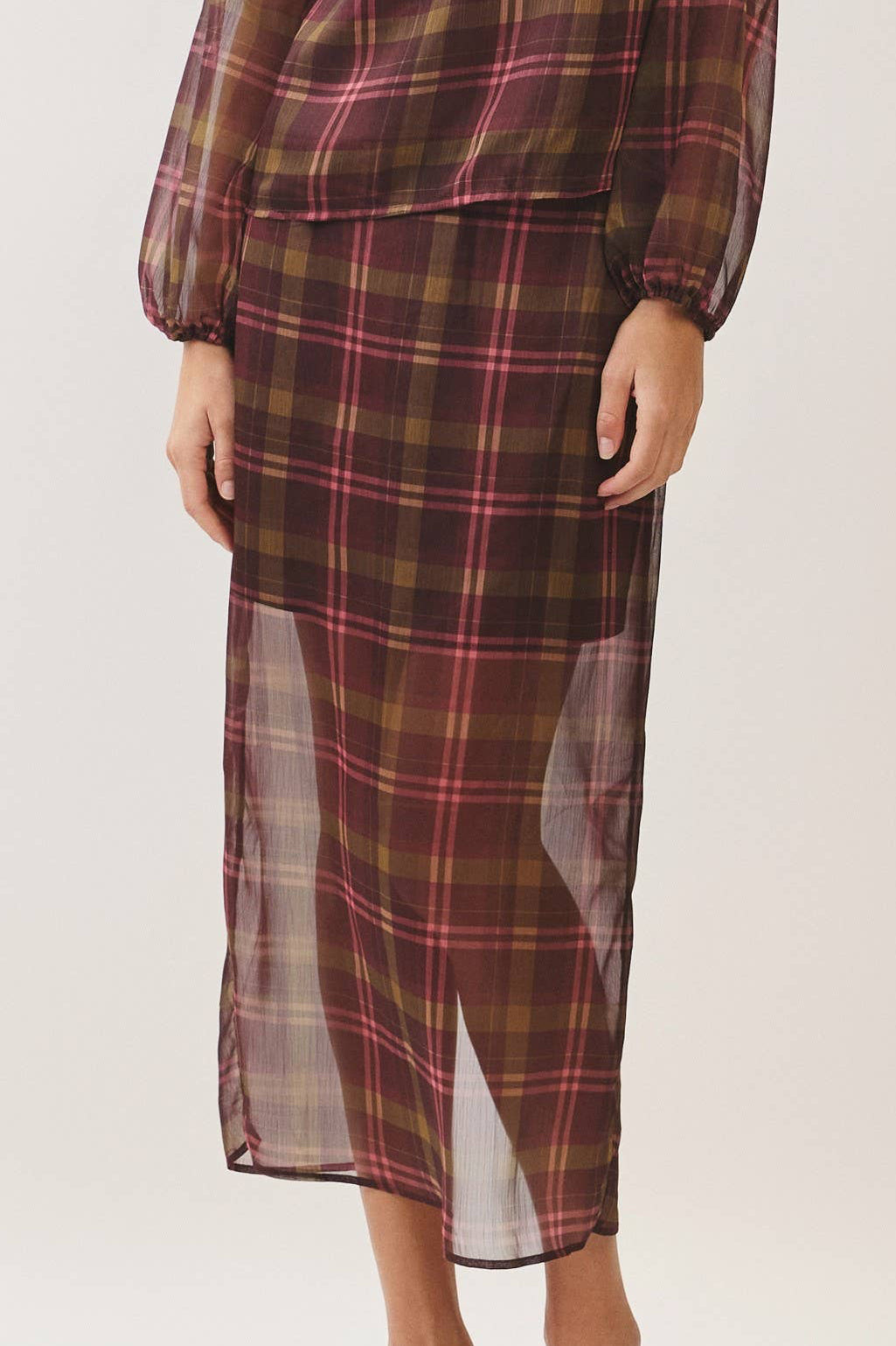 A woman modeling a matching plaid long-sleeve blouse and midi skirt set in a studio setting. The fabric features a brown, red, and black tartan pattern with sheer side panels.