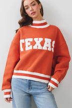 A woman wearing a burnt orange crewneck sweatshirt with 'TEXAS' printed in white across the chest, paired with light blue jeans, against a light gray studio background.