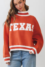 A woman modeling a rust-orange crewneck sweater with 'TEXAS' in white varsity letters across the chest, paired with light-wash wide-leg jeans and white sneakers, against a light gray studio background.