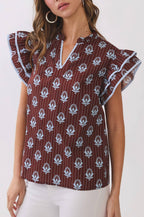 A woman modeling a short-sleeve blouse with flutter sleeves, featuring a brown base with white floral block prints and thin vertical stripes, paired with white pants against a plain white studio background.