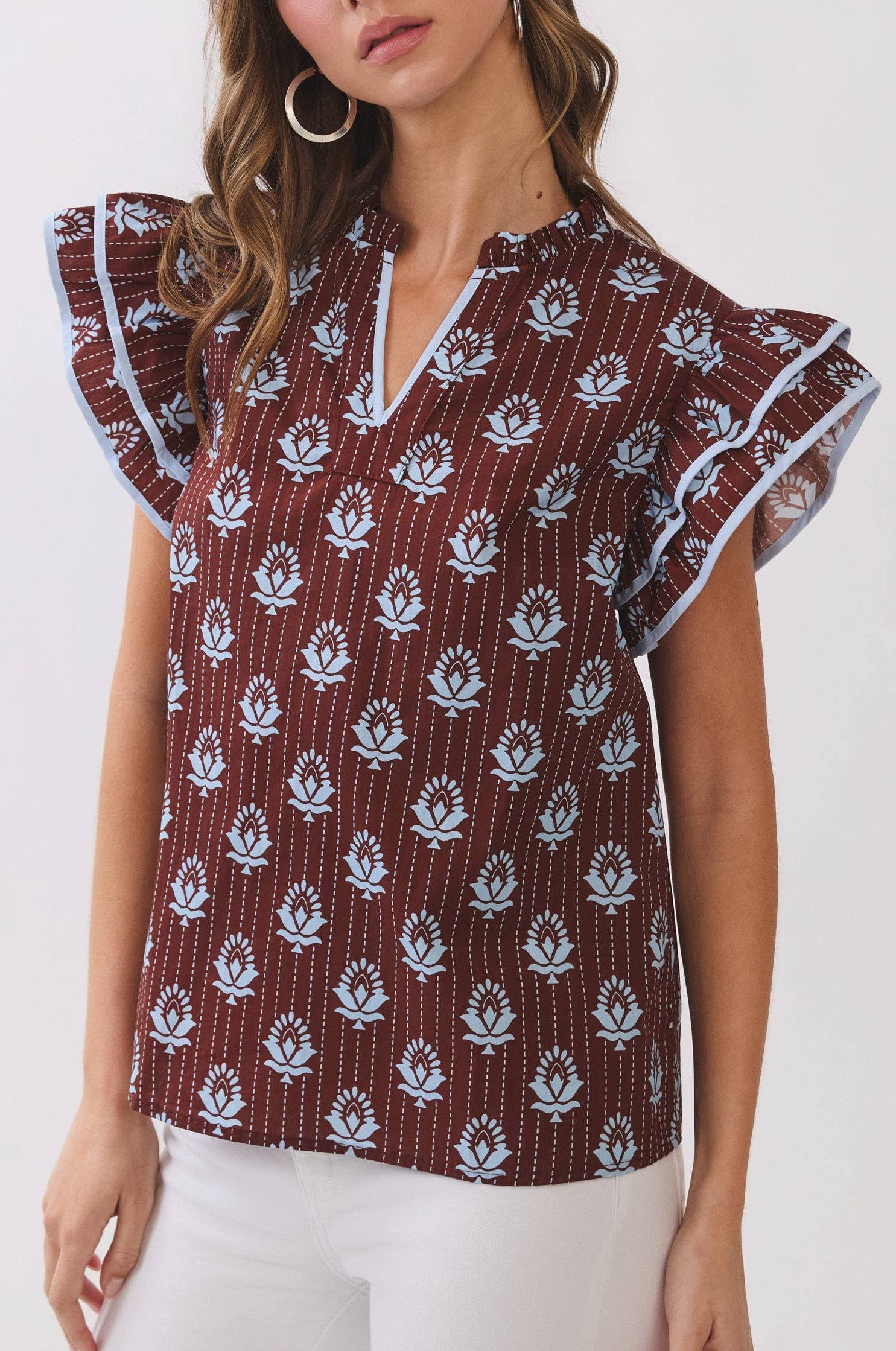 A woman modeling a short-sleeve blouse with flutter sleeves, featuring a brown base with white floral block prints and thin vertical stripes, paired with white pants against a plain white studio background.