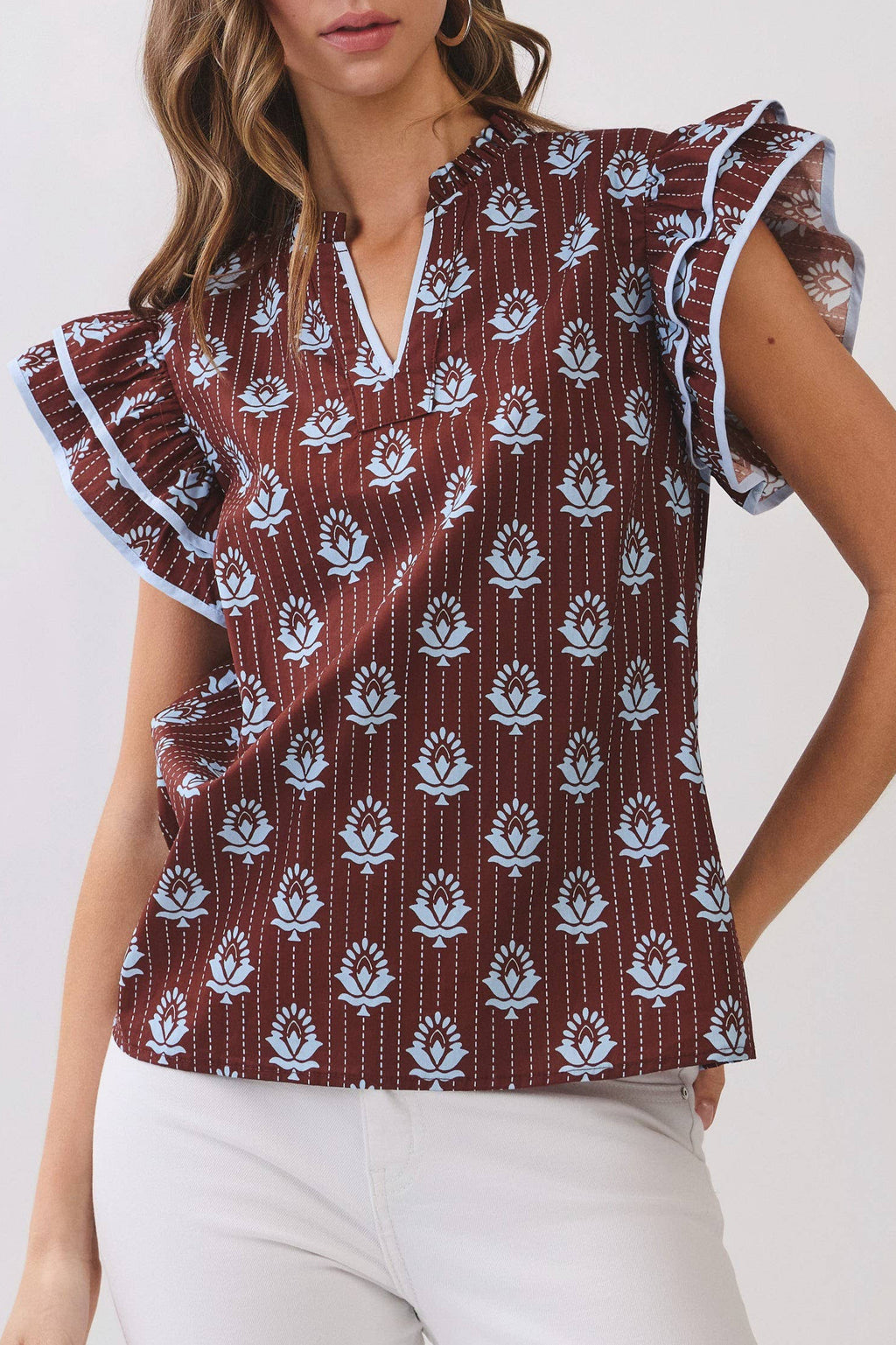 A woman modeling a short-sleeve blouse with flutter sleeves, a split V-neckline, and an allover white floral block print on a dark reddish-brown base, paired with white pants against a light gray studio background.