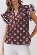 A woman modeling a short-sleeve blouse with flutter sleeves, a split V-neckline, and an allover white floral block print on a dark reddish-brown base, paired with white pants against a light gray studio background.