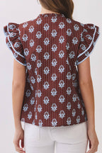 A woman viewed from the back wearing a brown ruffled blouse with a white floral block print and white pants, set against a plain off-white background.