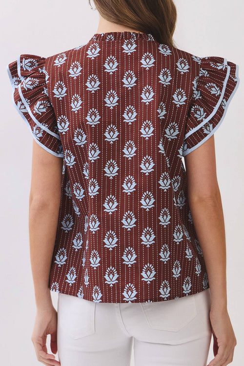 A woman viewed from the back wearing a brown ruffled blouse with a white floral block print and white pants, set against a plain off-white background.