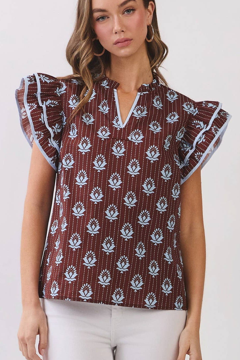 A woman modeling a short-sleeve top with flutter sleeves, featuring a brown base with a white floral ikat pattern, paired with white pants and beige sandals, set against a plain light gray studio background.