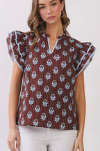 A woman modeling a short-sleeve top with flutter sleeves, featuring a brown base with a white floral ikat pattern, paired with white pants and beige sandals, set against a plain light gray studio background.
