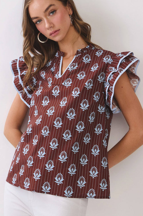 A woman wearing a brown short-sleeve top with a white floral block print and ruffled sleeves, paired with white pants, against a plain light gray background.
