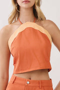 A woman modeling a matching two-piece outfit consisting of a halter crop top and a high-waisted mini skirt in orange with light beige contrast trim, set against a plain light gray studio background.