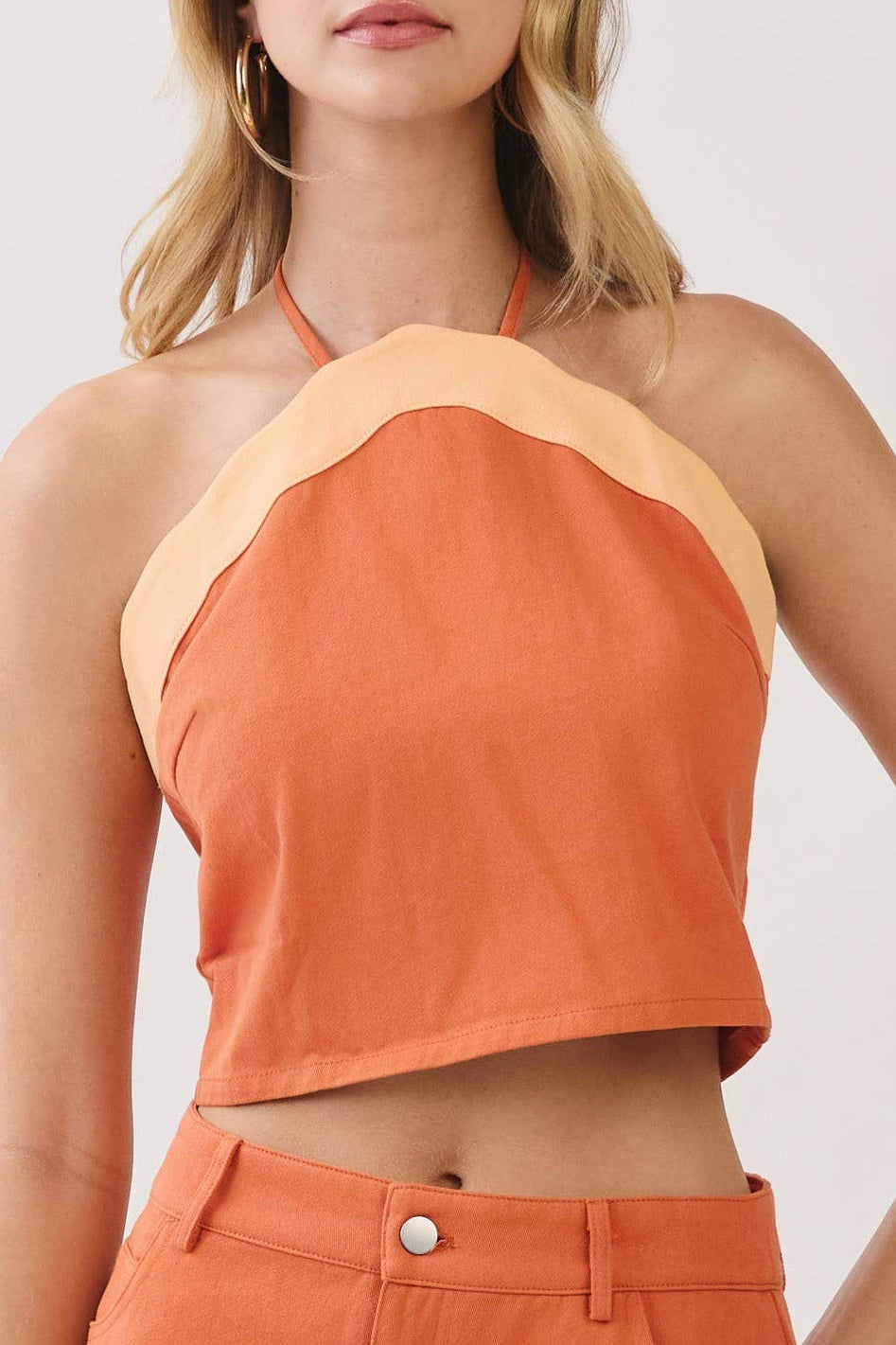 A woman modeling a matching two-piece outfit consisting of a halter crop top and a high-waisted mini skirt in orange with light beige contrast trim, set against a plain light gray studio background.