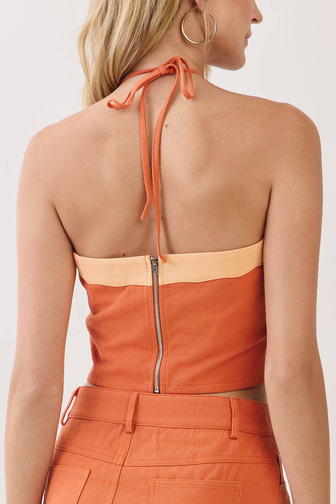 A woman shown from the back wearing a bright orange halter mini dress with beige contrast trim and a central zipper, photographed against a plain light background.