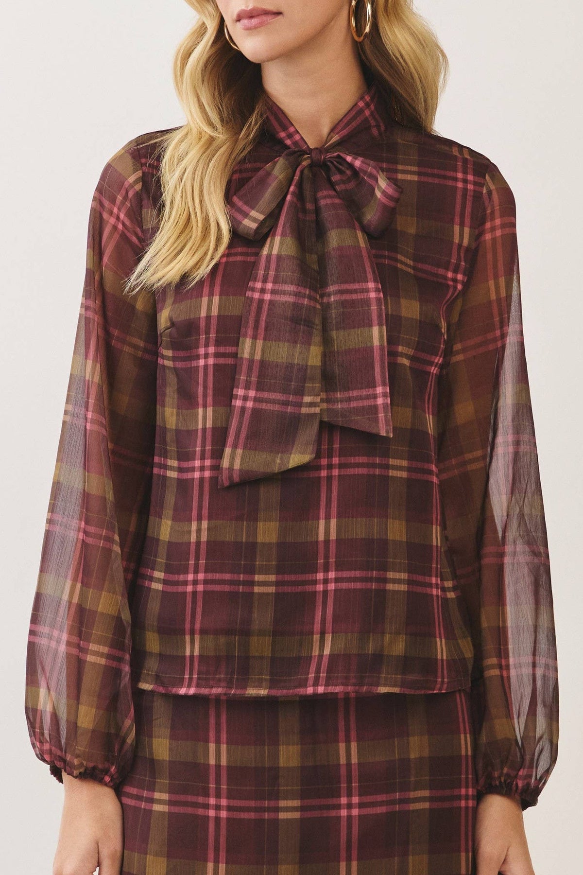 A woman modeling a matching plaid blouse and skirt set. The long-sleeve blouse features a sheer mesh overlay, a high collar, and a large bow at the neckline.
