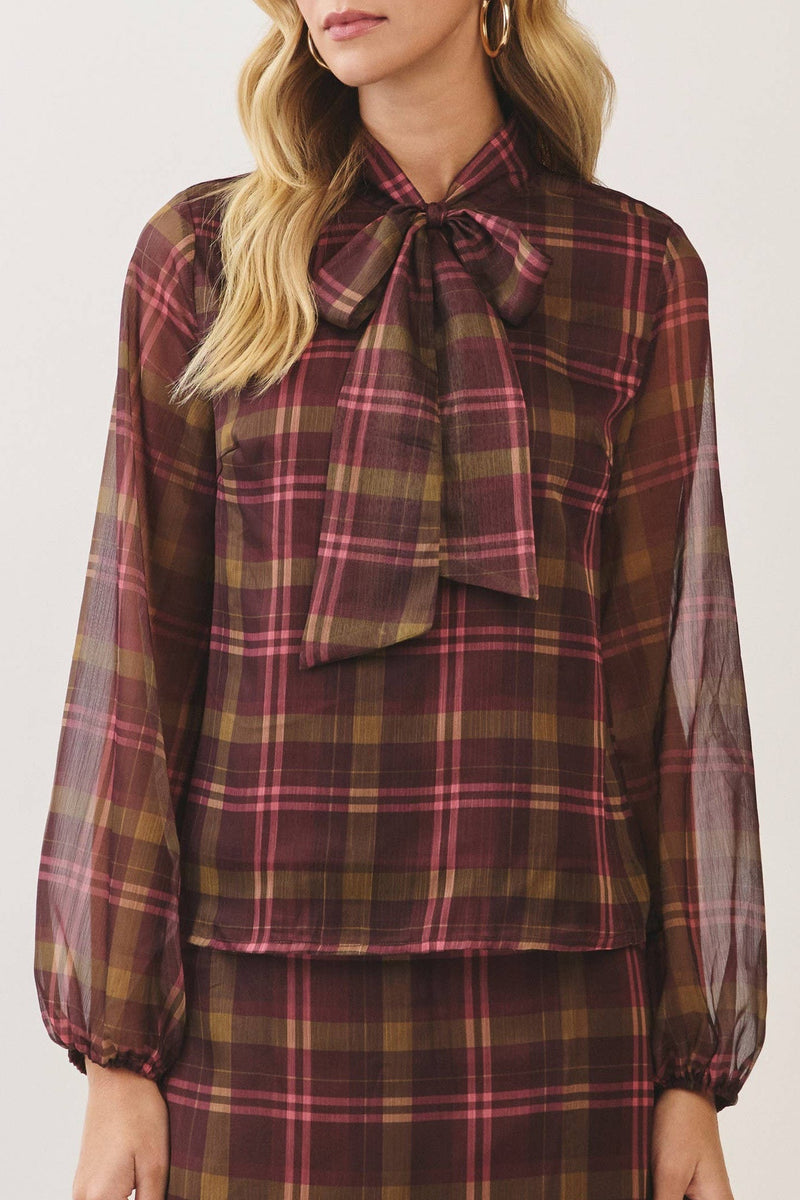 A woman modeling a matching plaid blouse and skirt set. The long-sleeve blouse features a sheer mesh overlay, a high collar, and a large bow at the neckline.