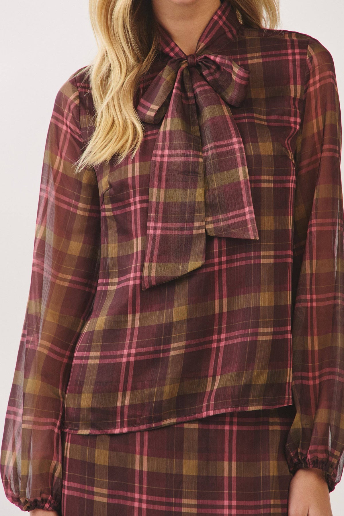 A woman wearing a sheer long-sleeve blouse with a large bow at the collar in a burgundy, pink, and olive green plaid pattern.