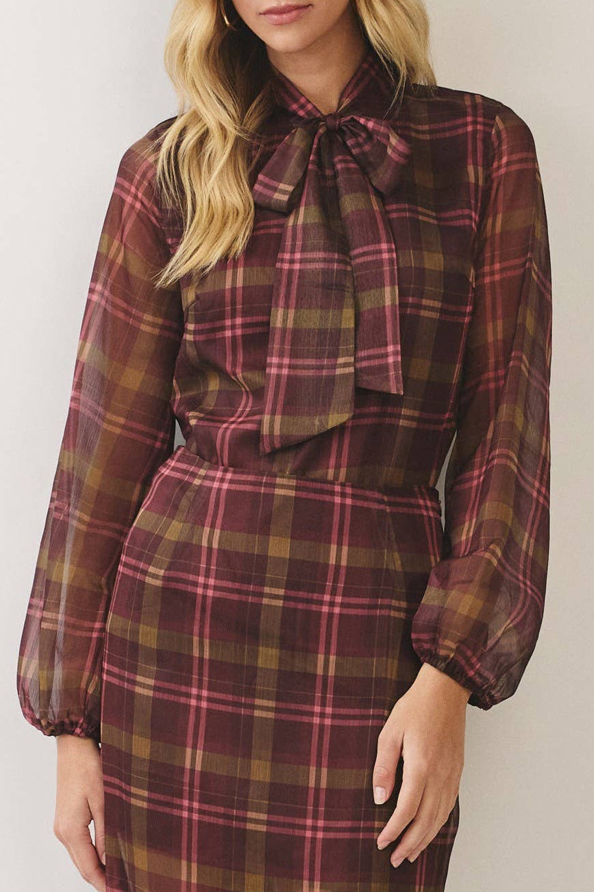 A woman modeling a long-sleeve midi dress with a brown, red, and black plaid pattern, featuring a ruffled tie-neck detail and sheer fabric, set against a plain light beige studio background.
