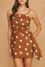 A woman modeling a brown mini dress with large white polka dots and thin spaghetti straps, standing against a textured tan background.