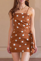 A woman modeling a brown strapless mini dress with white polka dots, standing in a warm-toned indoor setting with a tiled floor.