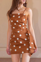 A woman modeling a brown mini dress with white polka dots, standing on a terracotta tiled floor against a beige wall.