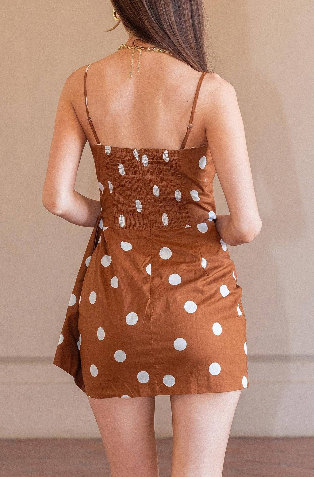 A woman viewed from behind wearing a brown mini dress with white polka dots and nude heels, standing on a terracotta tiled floor against a beige wall.