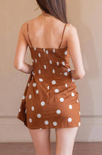 A woman viewed from behind wearing a brown mini dress with white polka dots and nude heels, standing on a terracotta tiled floor against a beige wall.