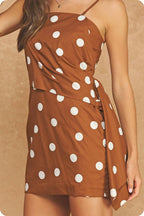 A woman wearing a brown wrap-style mini dress with large white polka dots, shown from the shoulders to the upper thighs against a warm beige textured background.