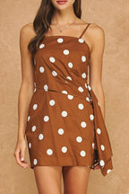 A woman modeling a brown mini dress with white polka dots, featuring thin spaghetti straps and a wrap-style skirt, set against a textured tan studio backdrop.