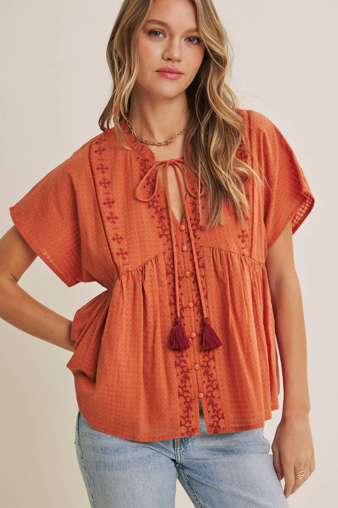 Woman wearing an orange embroidered top with tassels on a beige background