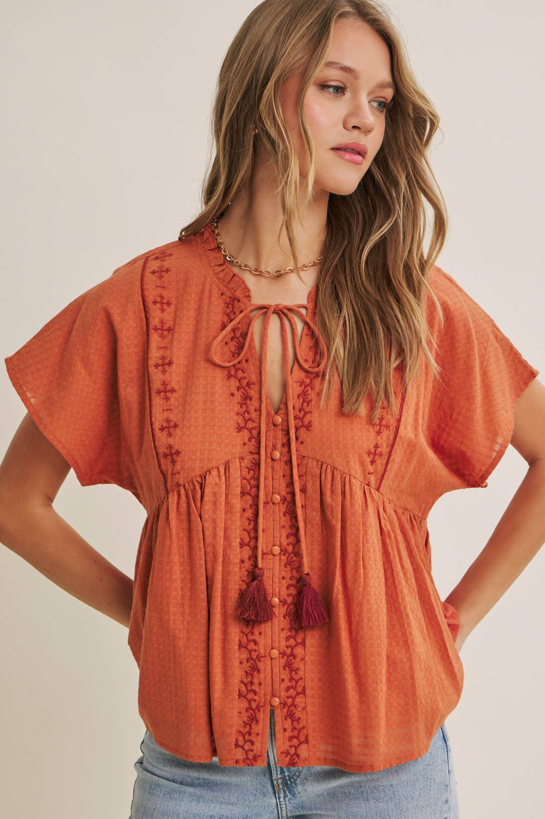 Woman wearing an orange embroidered blouse with tassels on a plain background