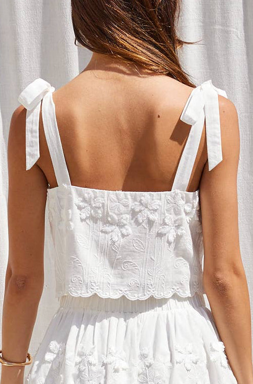 A woman shown from the back wearing a white sleeveless top with floral embroidery and bow straps, standing against a white curtain backdrop.