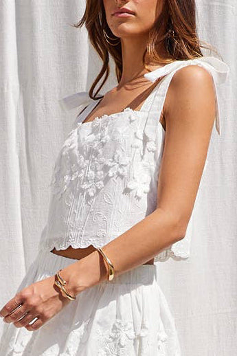 A woman modeling a white sleeveless lace-trimmed top with a ruffled hem, paired with beige strappy platform sandals, posed in front of a white draped fabric backdrop.
