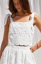 A woman modeling a white two-piece outfit consisting of a sleeveless crop top and matching flared skirt, paired with beige lace-up platform sandals, set against a draped white fabric backdrop.