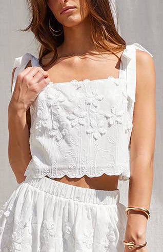 A woman models a white two-piece outfit consisting of a sleeveless crop top with floral embroidery and a matching flared skirt, paired with beige lace-up platform sandals, set against a white draped fabric backdrop.